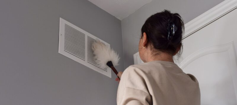 woman dusting dirty air vent on the gray wall of her home